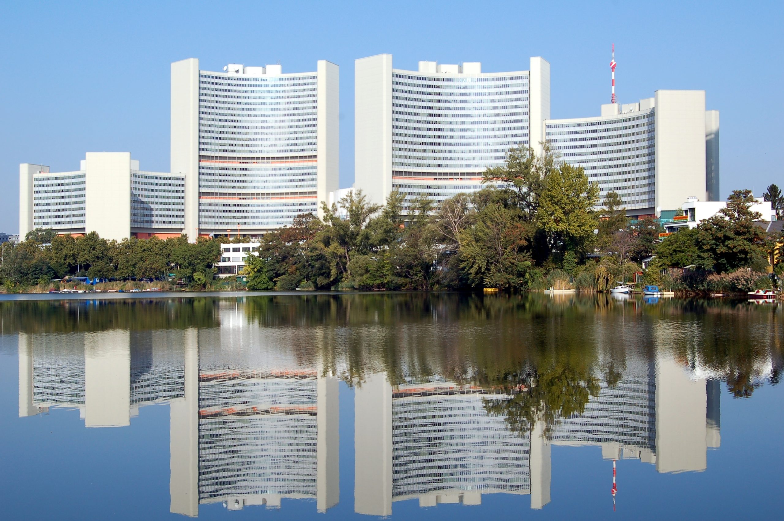 Die weißen hohen Gebäude der Uno City spiegeln sich im Kaiserwasser wieder, umgeben von Bäumen unter einem klaren blauen Himmel.
