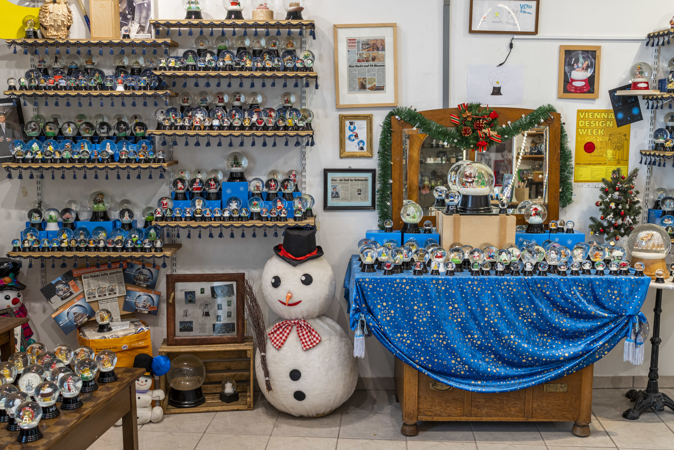 Raum voller Schneekugeln der Wiener Schneekugelmanufaktur auf Regalen, eine große Schneemannfigur und festliche Dekoration auf einer blauen Tischdecke.