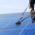 Eine Person reinigt an einem sonnigen Tag Solarmodule mit einer langen Bürste und Wasser.