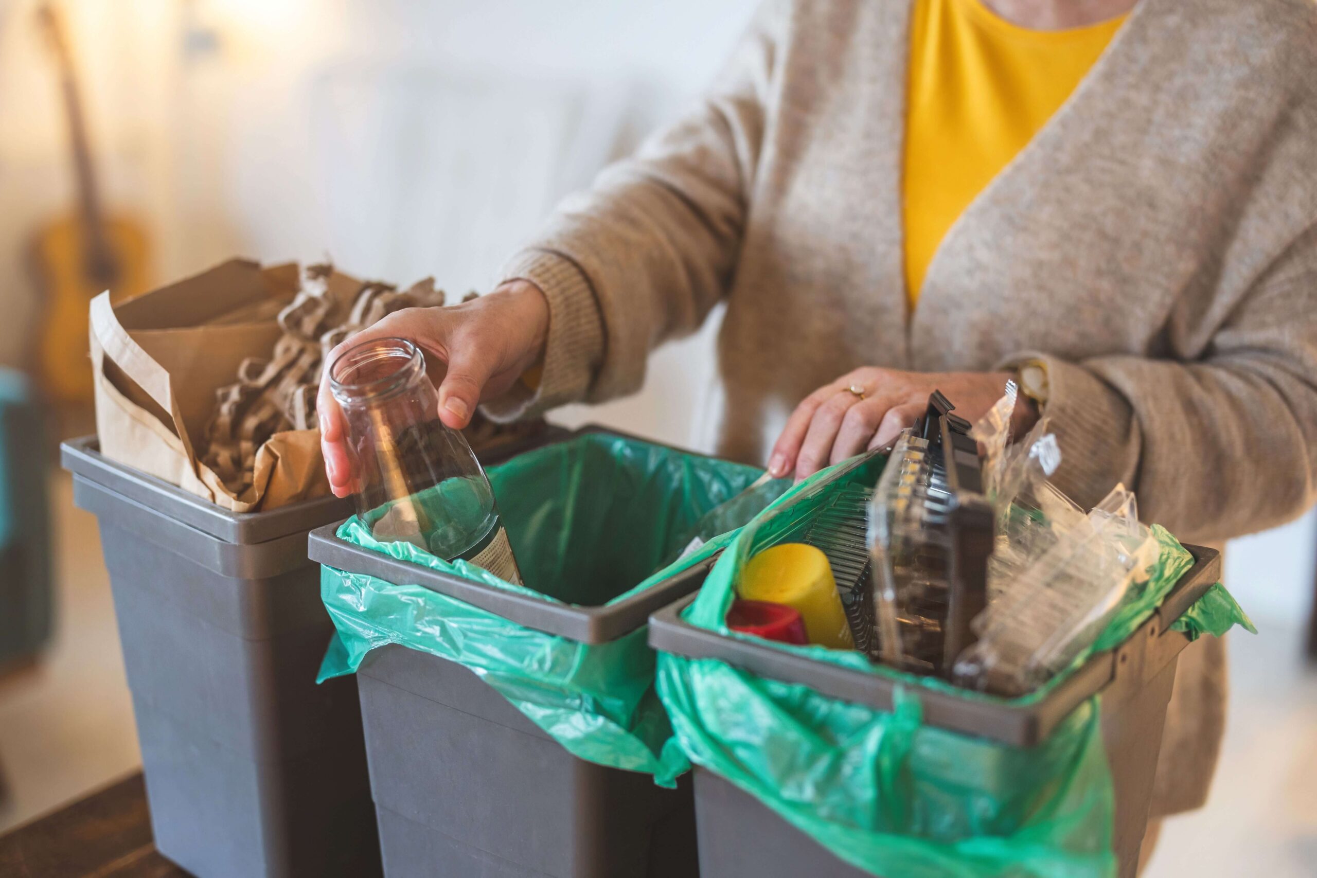 Person sortiert Wertstoffe in Behälter und hält ein Glasgefäß in der Hand, in dem Papier- und Plastikgegenstände sichtbar sind.